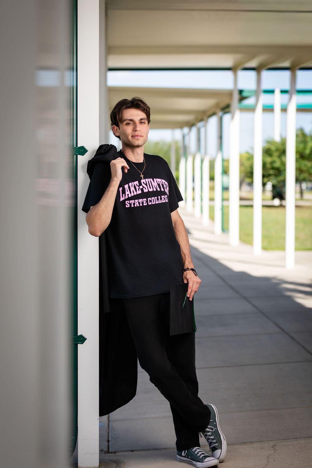 Young man in black Lake-Sumter State College t-shirt and pants leans against column in outdoor corridor.