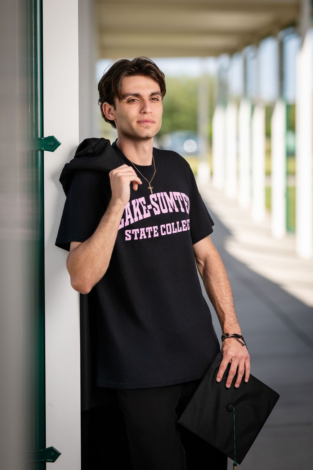 Young man in black Lake-Sumter State College t-shirt holds graduation cap while leaning against white column outdoors.
