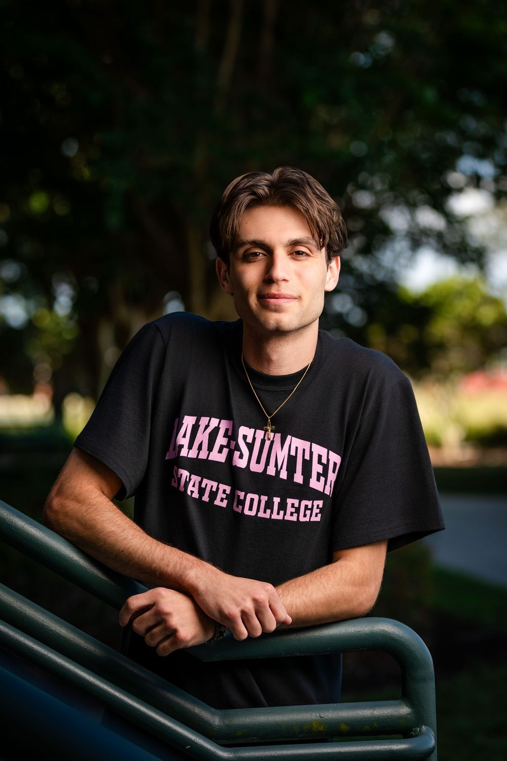 Young man in black Lake Sumter State College t-shirt leaning on railing outdoors, smiling confidently.