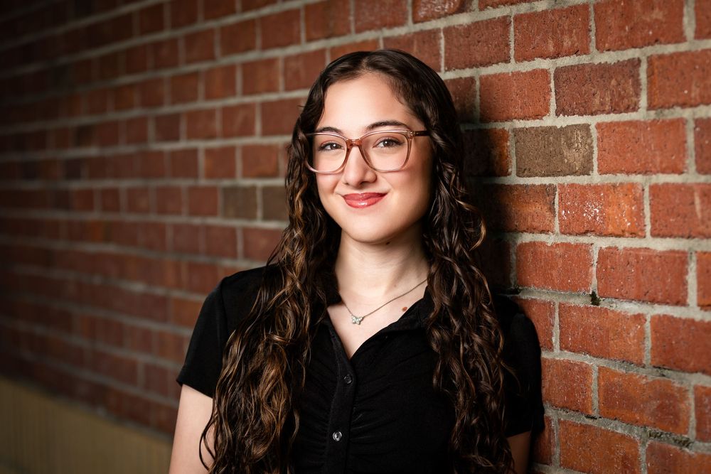 Young woman with curly hair and glasses smiling against a brick wall backdrop.