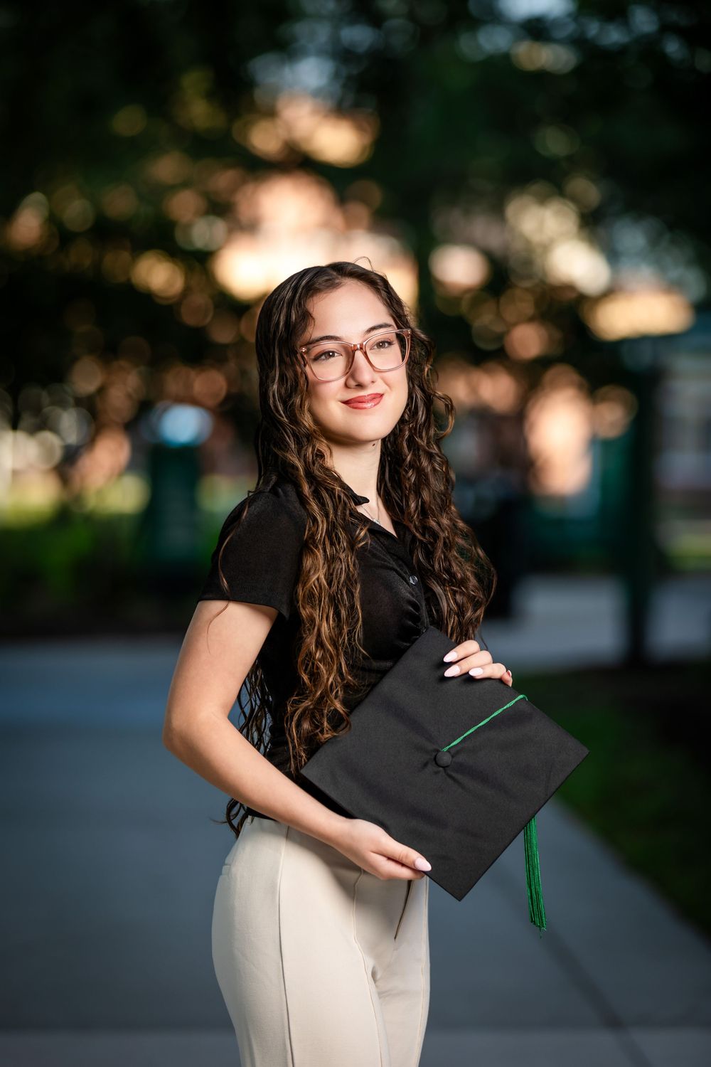 Young woman in glasses holding a black graduation cap with green tassel, smiling outdoors in soft evening light.