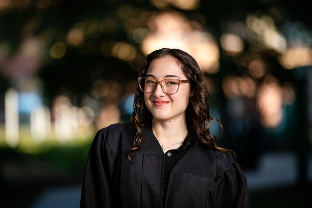 Smiling young woman in black graduation gown and glasses posing outdoors at dusk on campus.