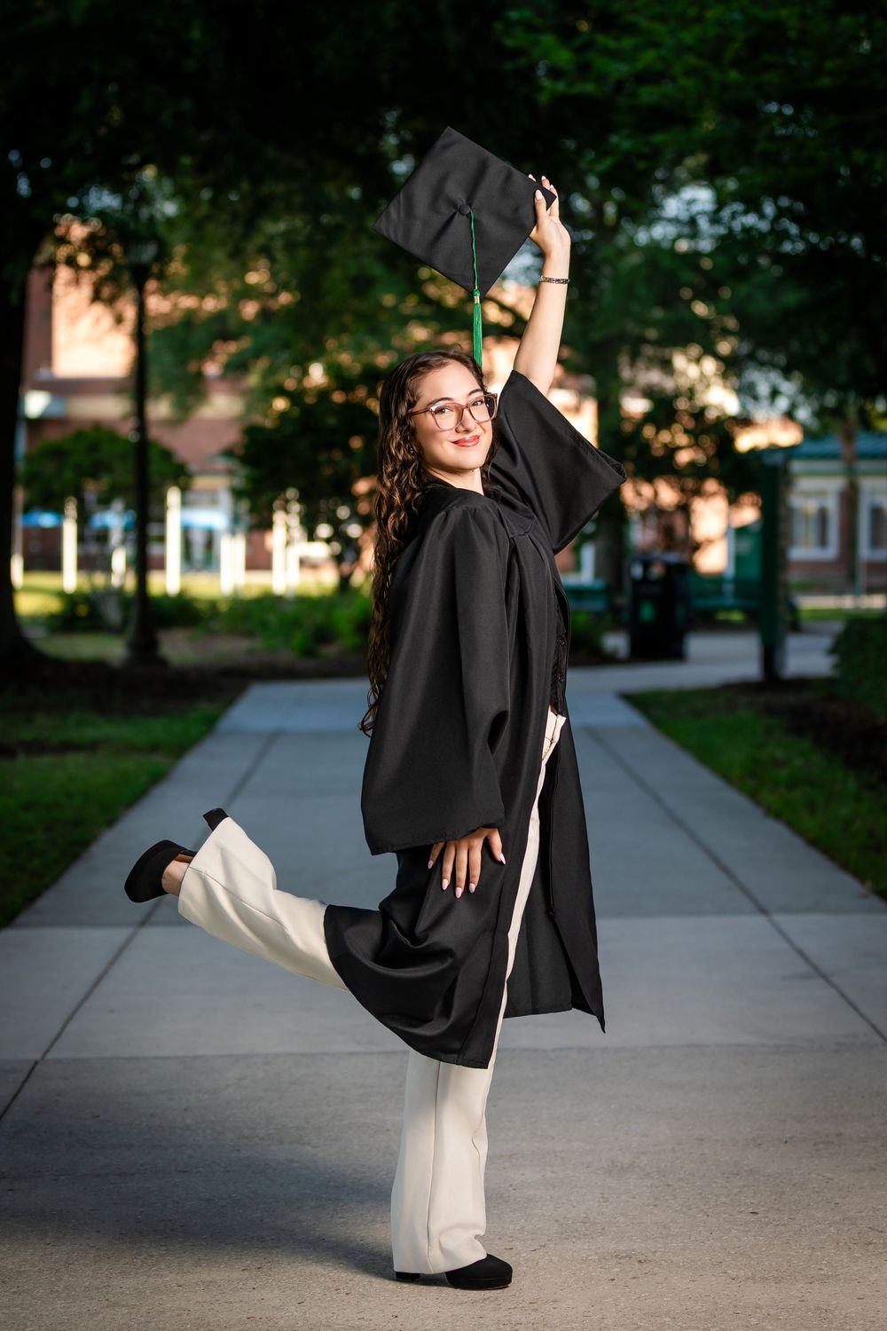 A joyful graduate in black cap and gown poses playfully on a campus pathway, lifting her cap and leg in celebration.