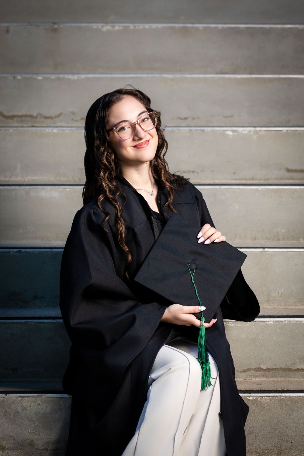 Young woman in black graduation gown and cap with green tassel smiling on steps.