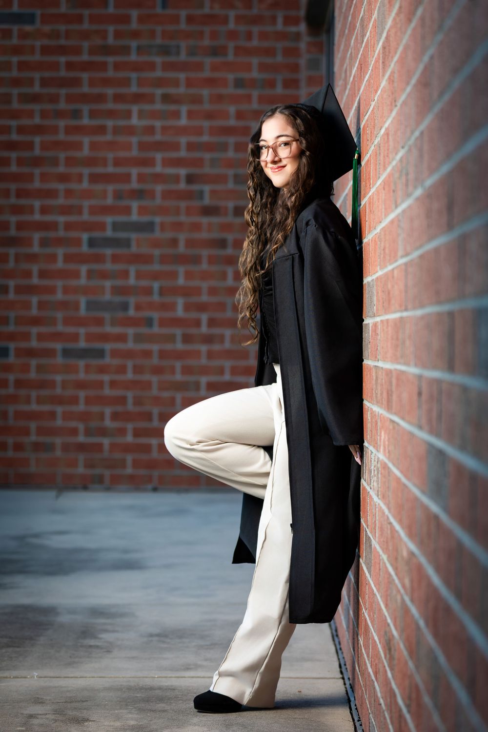 Young woman in black graduation gown and cap leans against brick wall, wearing glasses and white pants.