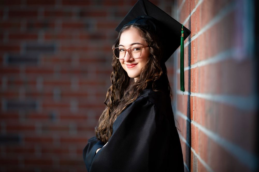 Young woman in graduation cap and gown smiling against brick wall, wearing glasses and academic regalia.
