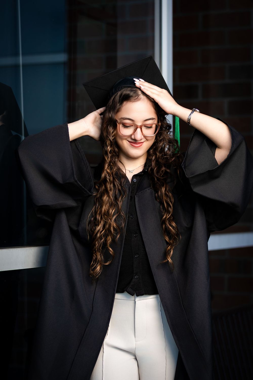 Young woman in black graduation gown and cap smiling, adjusting mortarboard, wearing glasses and white pants.