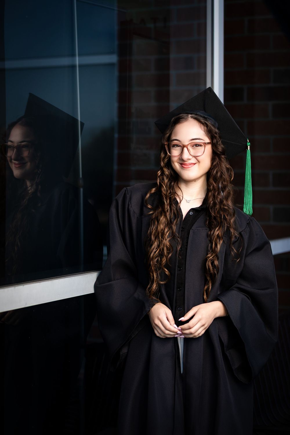 Young woman in black graduation cap and gown with green tassel smiles proudly, reflection visible in window behind her.