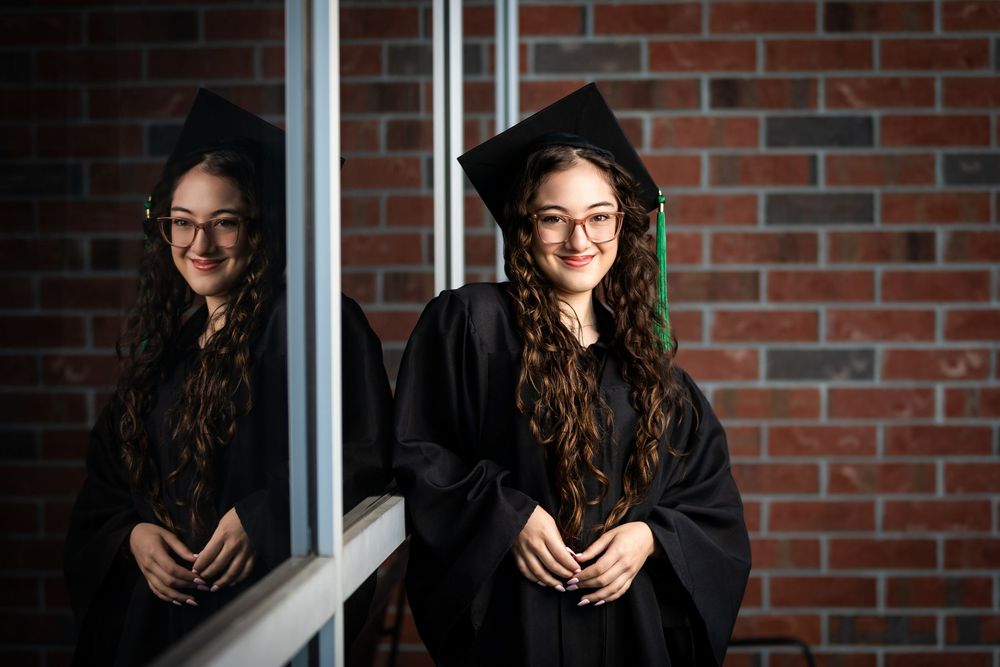 Young woman in black graduation gown and cap smiles beside her reflection in a mirror against a brick wall.
