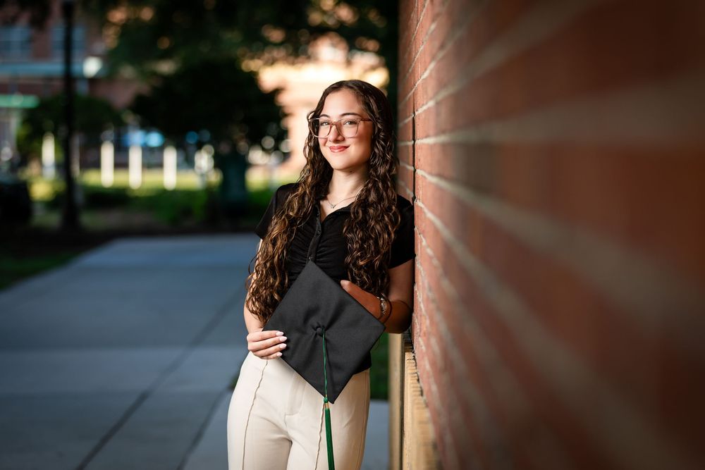 A young woman in glasses holds a graduation cap while leaning against a brick wall outdoors.