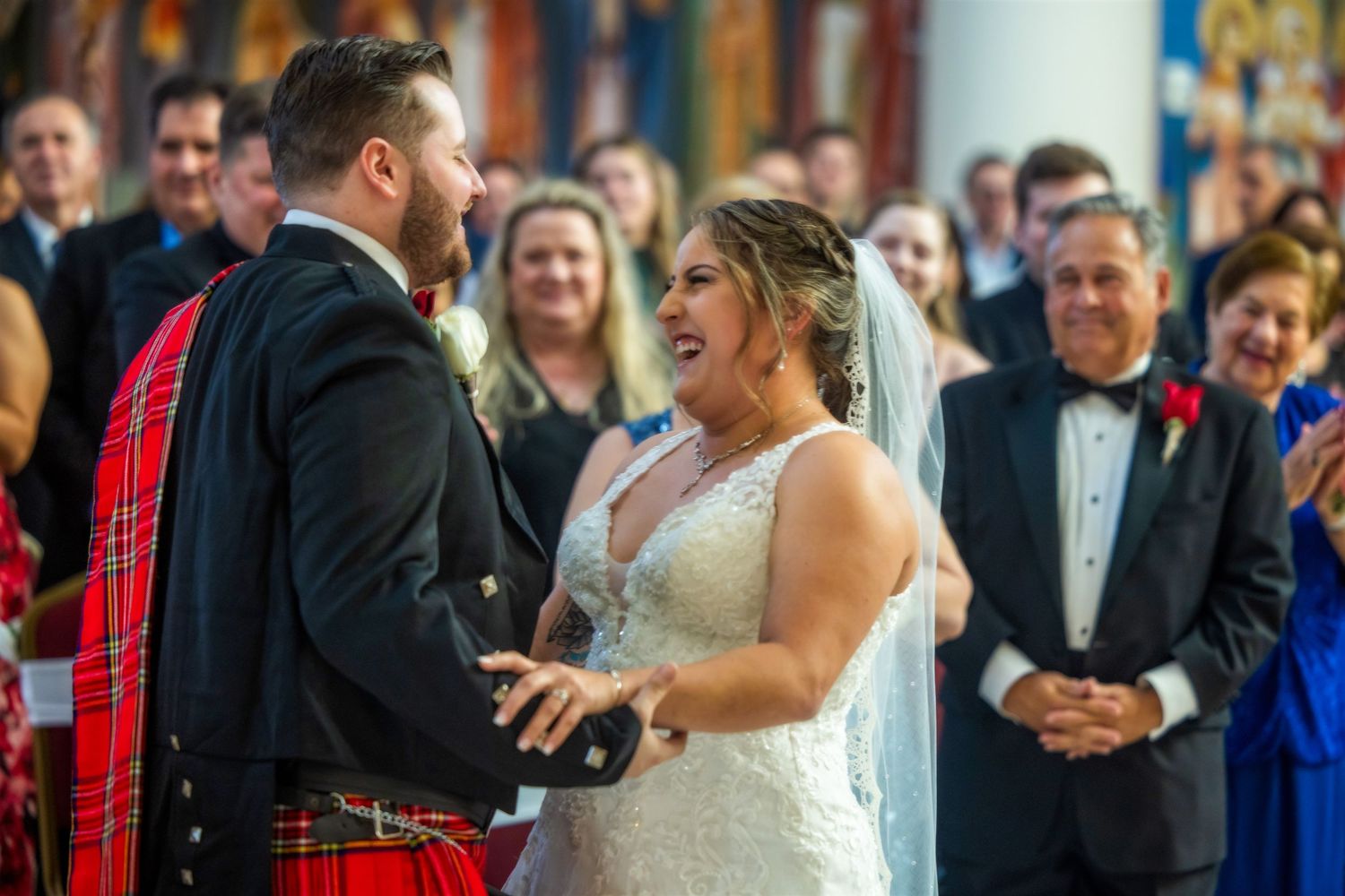 Bride and groom exchange vows at altar, groom wearing red Scottish kilt, bride in lace gown, guests smiling behind them.