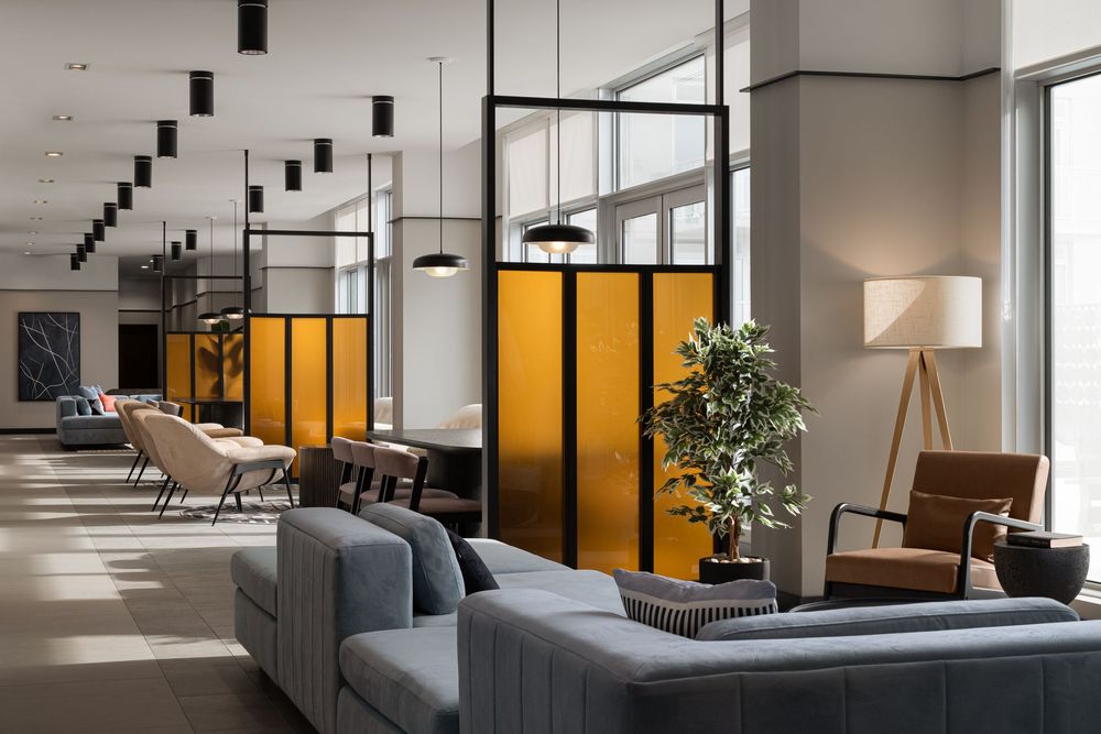 Modern hotel lobby with gray sofas, yellow partition panels, pendant lights, and a potted tree near large windows.