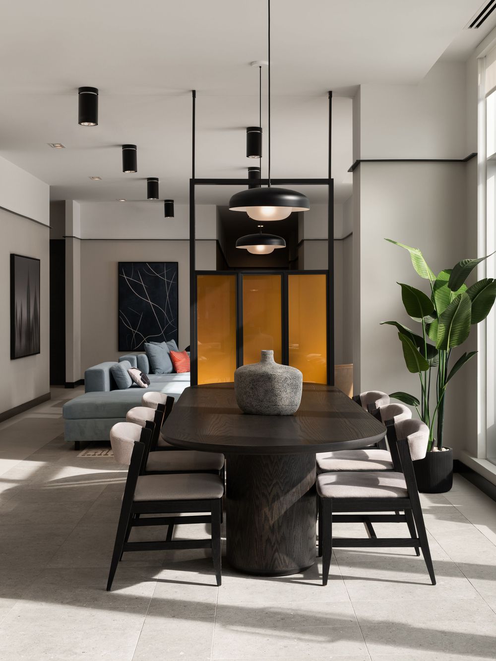 Modern dining room with dark oval table, black pendant lights, yellow accent wall, and tropical plant in neutral toned open plan space.
