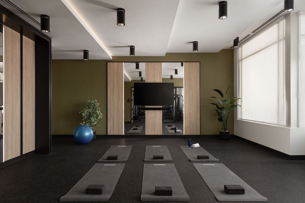 Modern yoga studio with gray mats, olive green walls, wooden cabinetry, and a blue exercise ball on dark flooring.