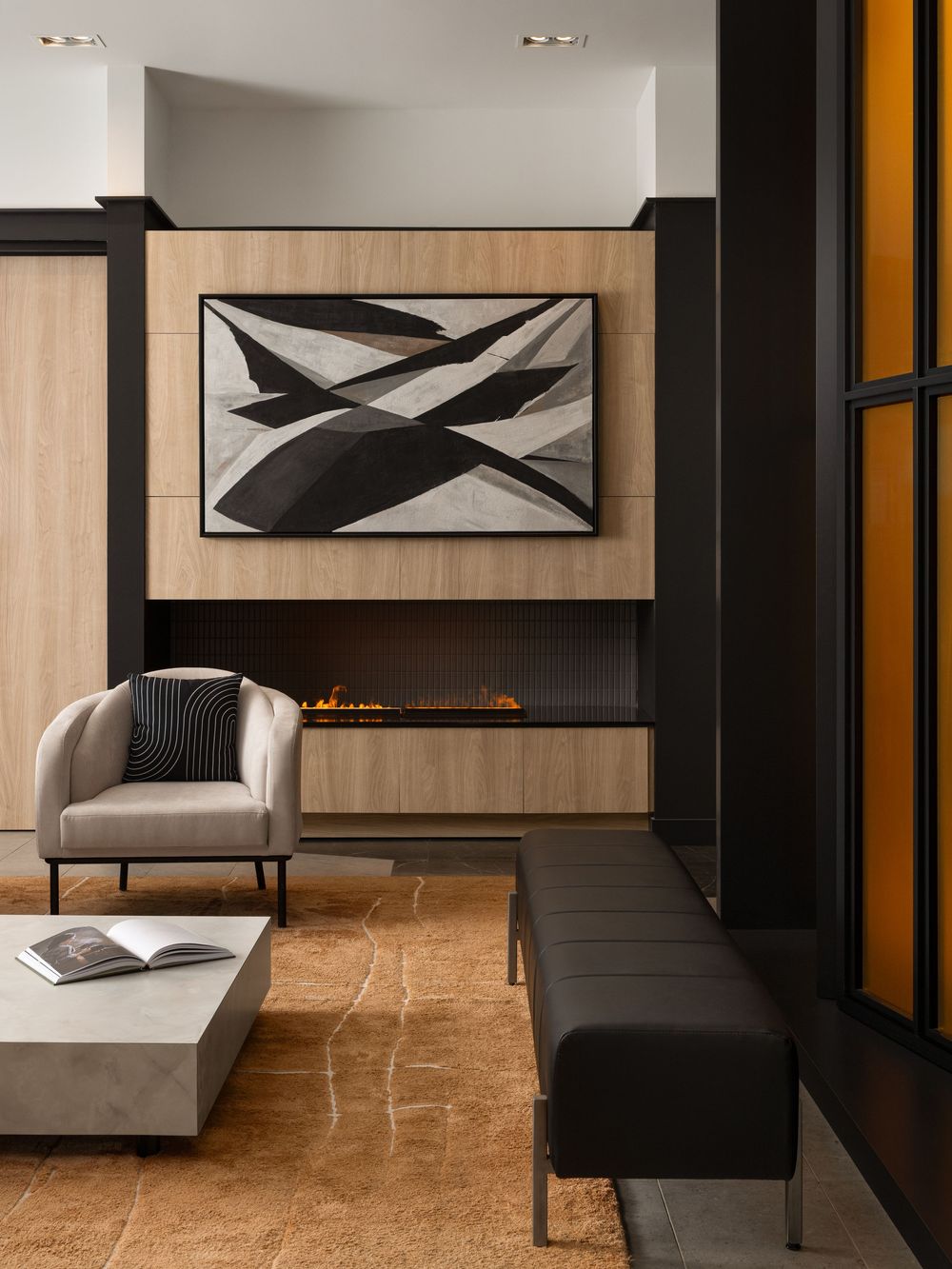Modern living room with linear fireplace, wood paneling, abstract art, gray armchair, marble coffee table, and dark bench seating.