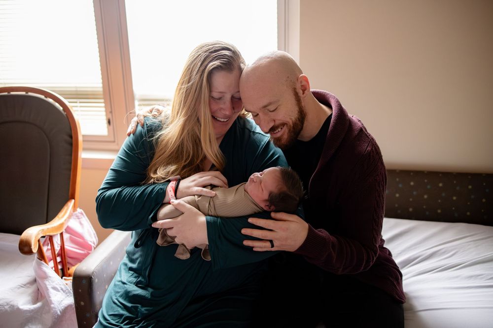 New parents lovingly cradle their newborn baby together on a bed near a bright window.