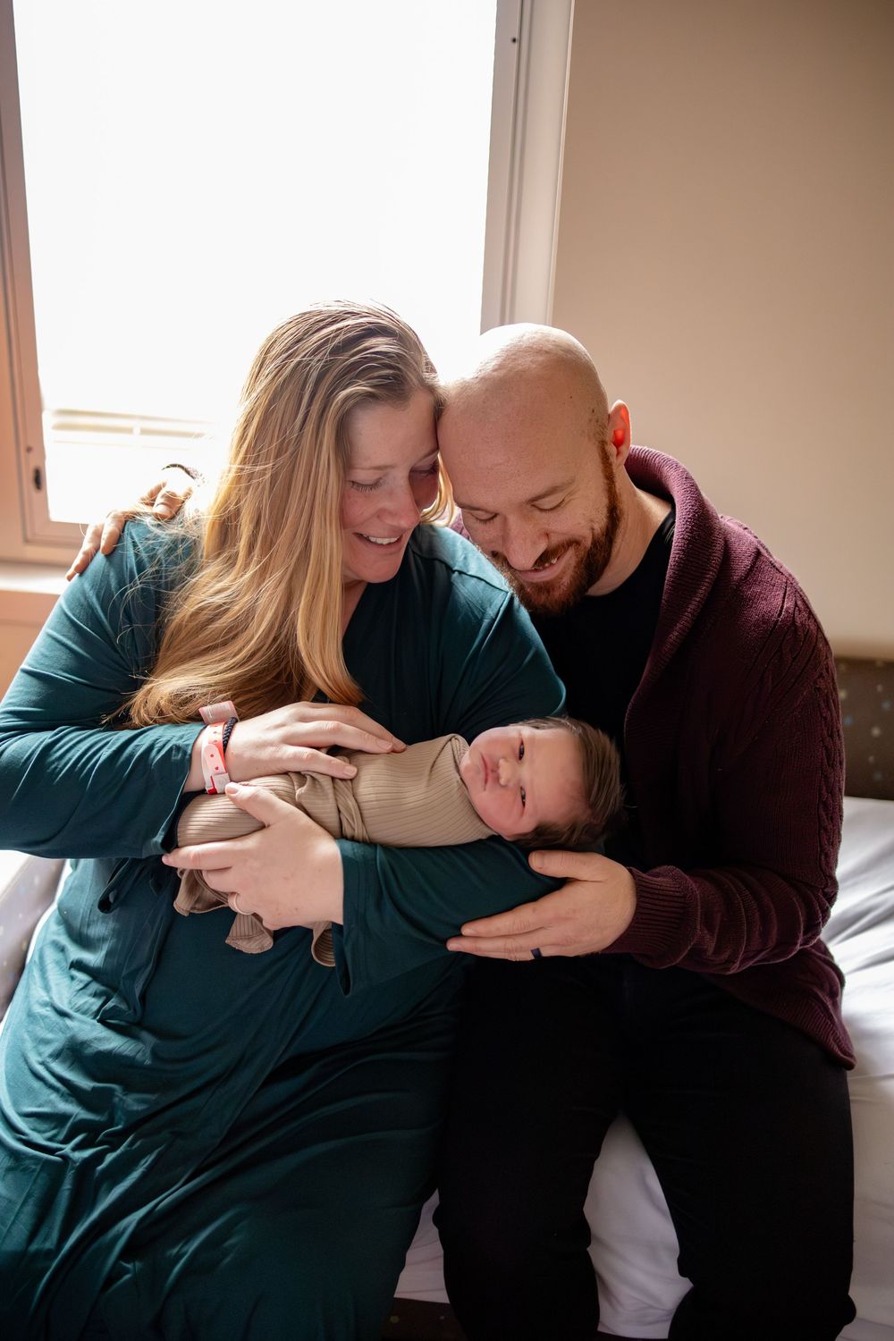 A couple smiles while cradling their swaddled newborn baby together, sitting by a bright window in a warm, intimate moment.