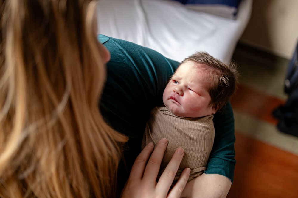 Mother cradling a swaddled newborn baby in a tan wrap in a hospital room.