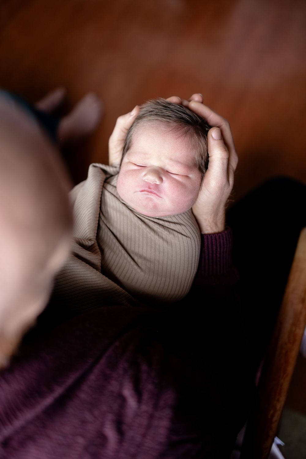 Newborn baby sleeping peacefully wrapped in beige, cradled gently in adult hands against warm brown background.