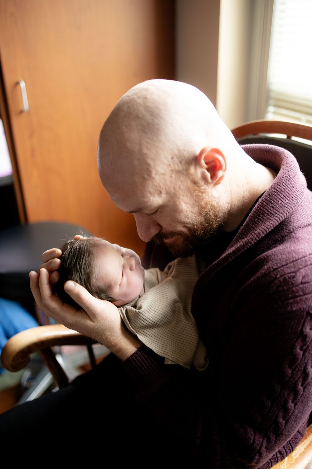 Bald bearded father tenderly holds sleeping newborn baby close to his face while seated in a wooden chair.