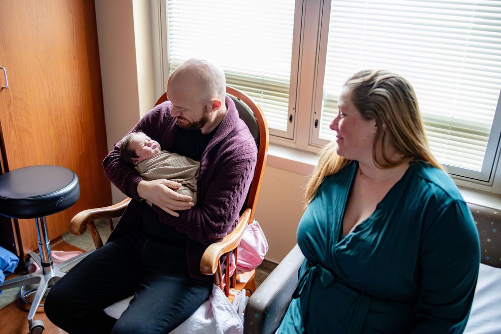 New parents admire their newborn baby, father cradling infant in chair while mother looks on lovingly.