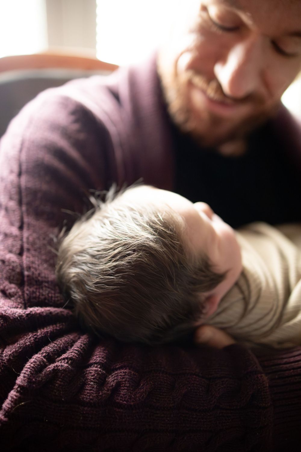Smiling bearded man in purple sweater tenderly holding a sleeping newborn baby in his arms.