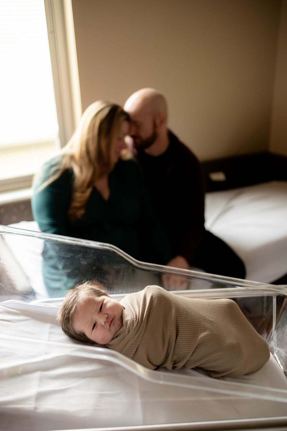 Newborn baby swaddled in hospital bassinet with proud parents sharing a tender moment in the background.