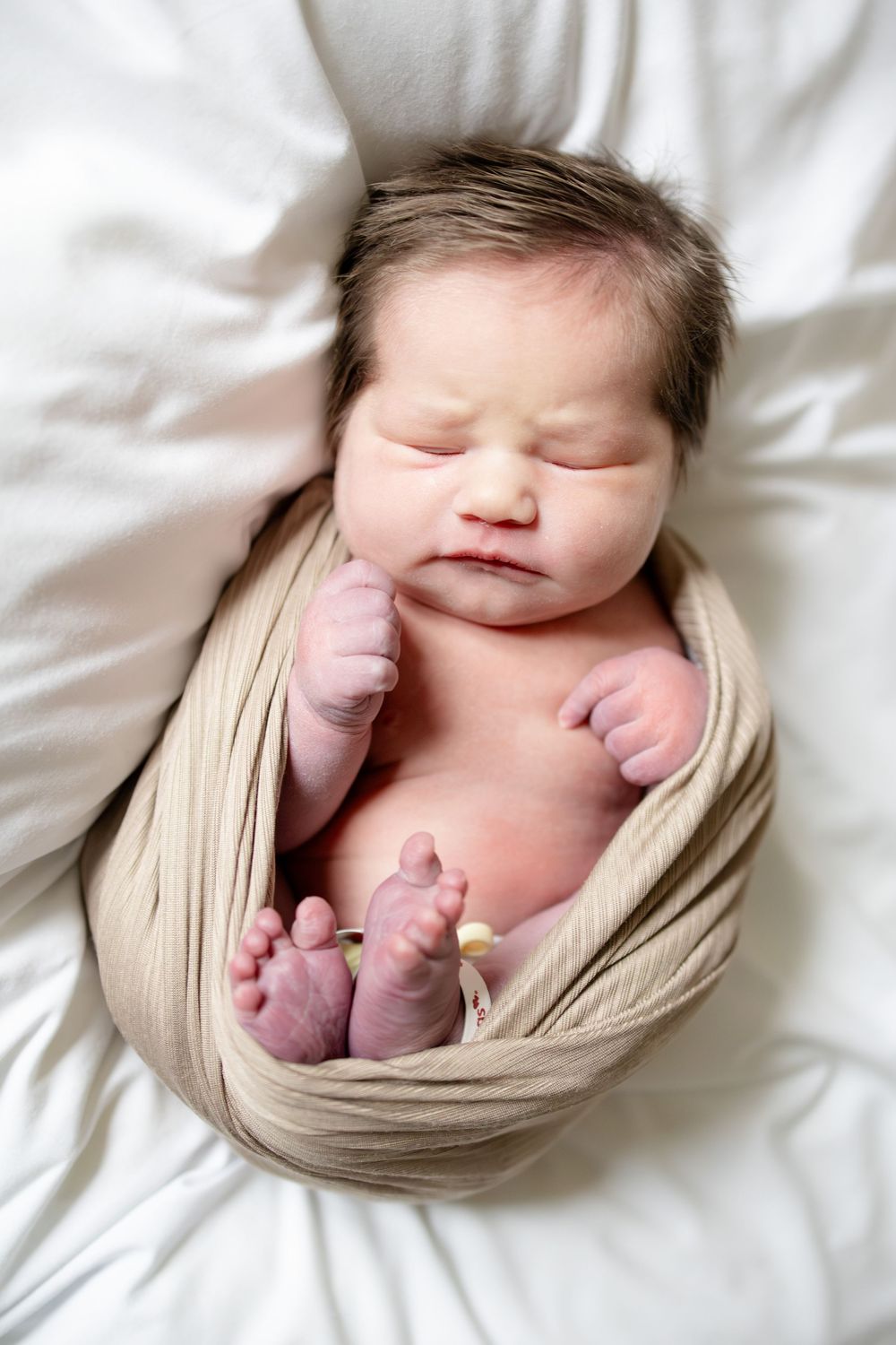 Newborn baby sleeping peacefully, swaddled in a beige wrap, with tiny hands and feet visible on white bedding.