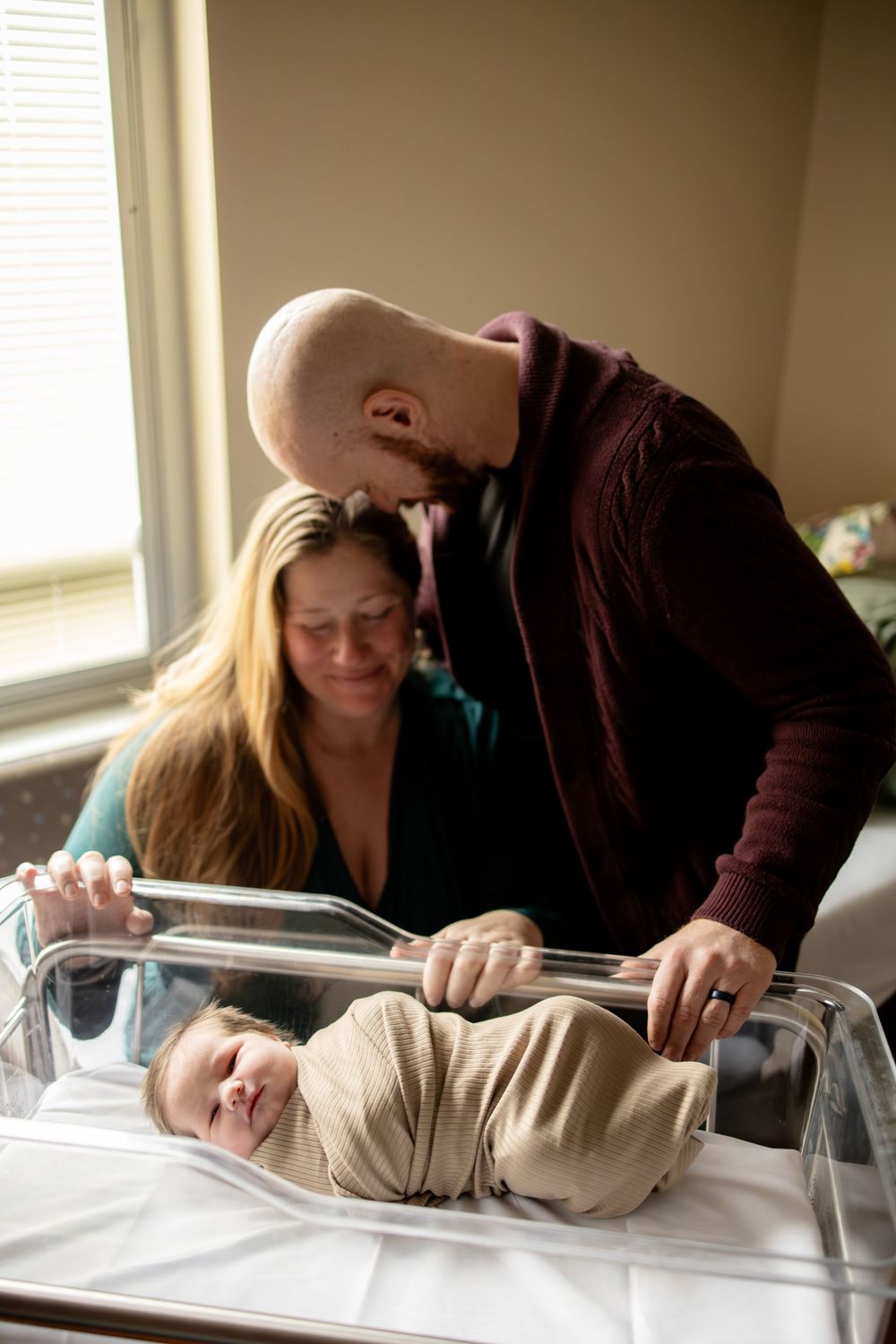New parents lean over hospital bassinet admiring their swaddled newborn baby together.