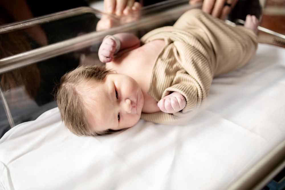 Newborn baby lying on white hospital blanket in bassinet, wearing beige onesie, with eyes open.