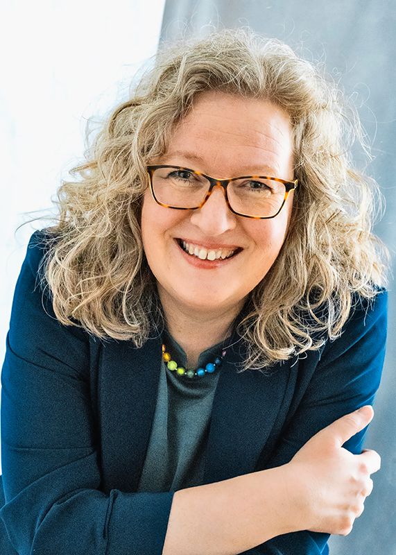 Smiling woman with curly blonde hair, glasses, and colorful necklace wearing a navy blazer in a professional portrait.