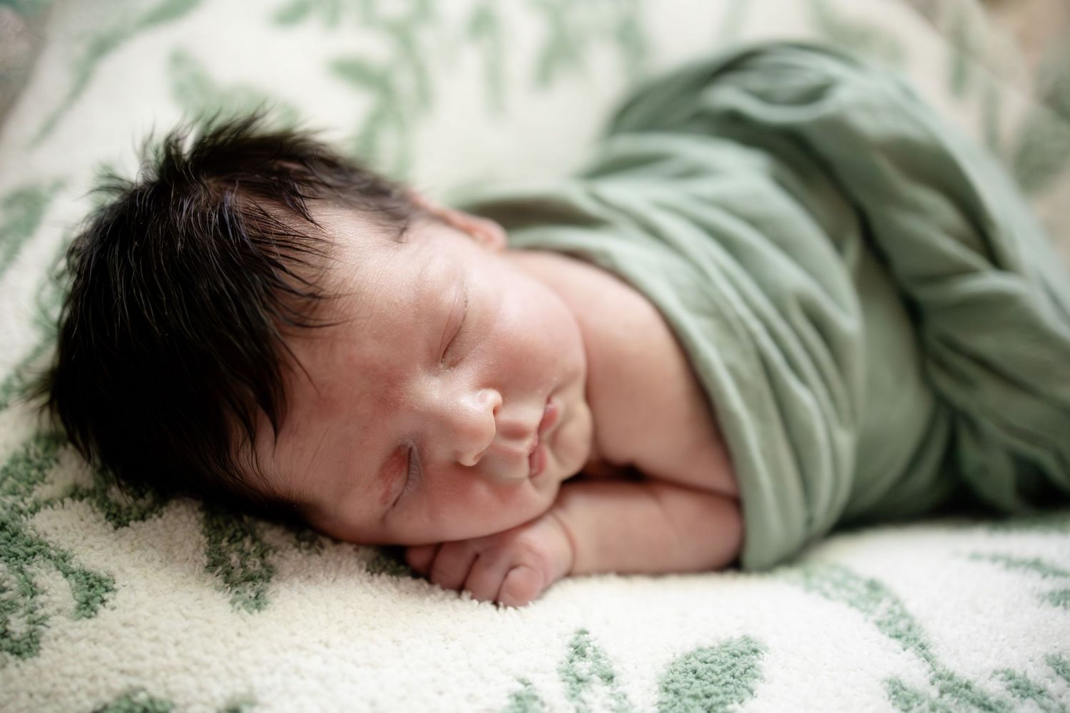 Peaceful newborn baby with dark hair sleeping wrapped in a green swaddle on a floral white blanket.
