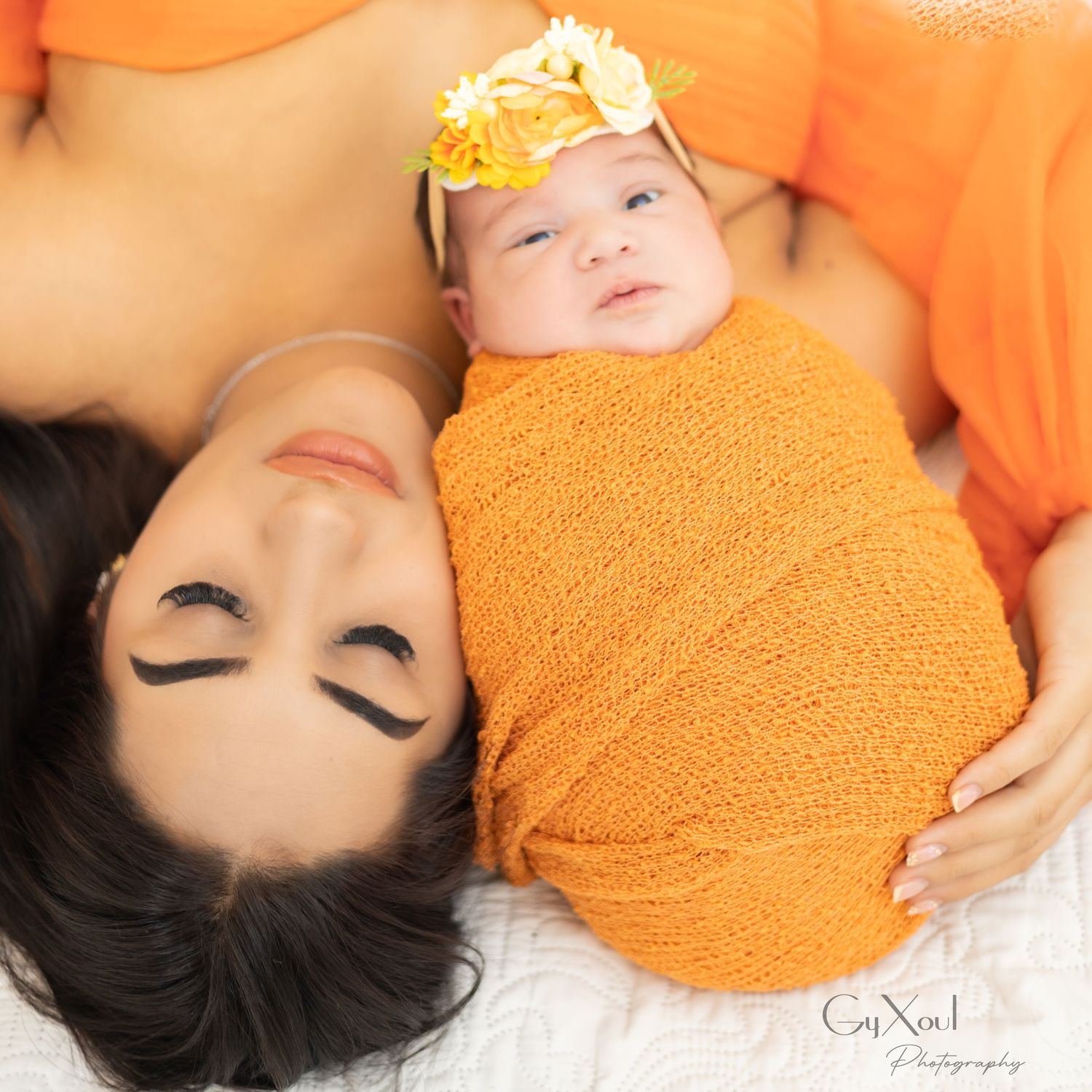 Mother in orange dress lying with newborn baby swaddled in orange wrap wearing yellow floral headband.