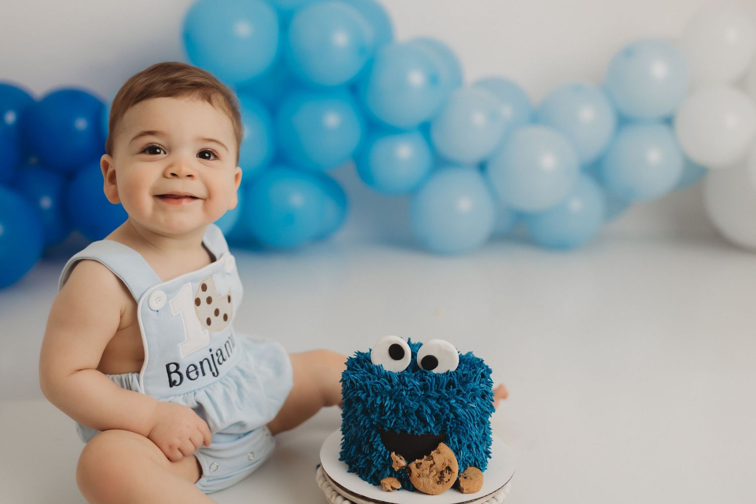 Baby boy in Cookie Monster romper smiles beside a blue furry smash cake, surrounded by blue balloons.