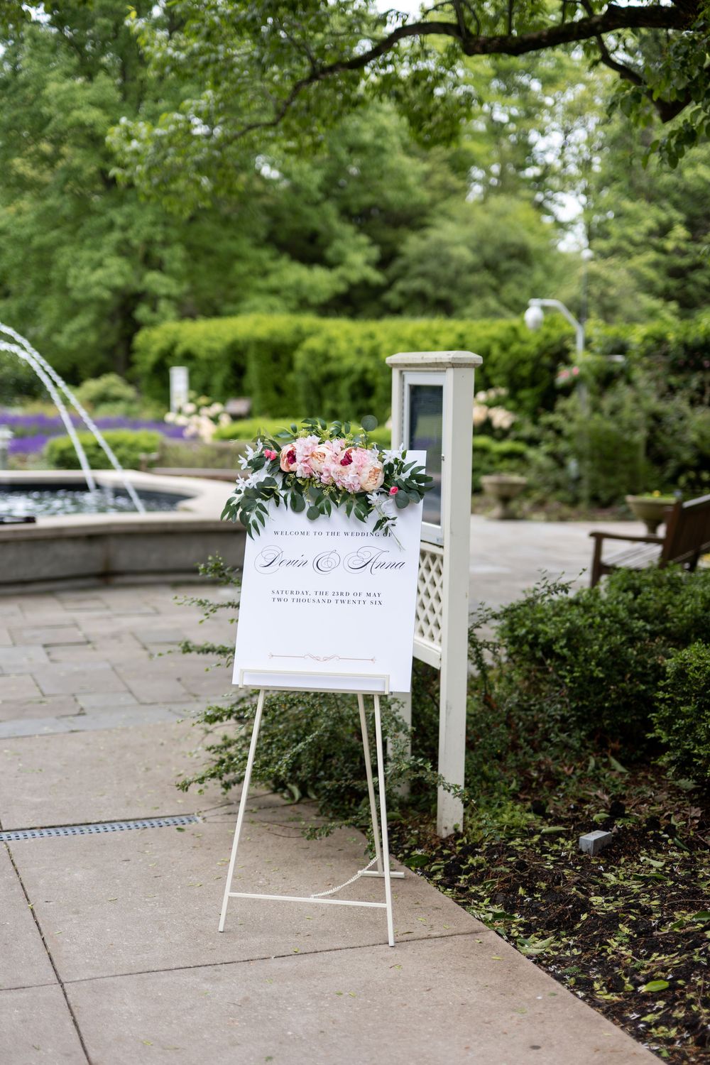 White wedding welcome sign with pink floral arrangement on easel in garden setting.