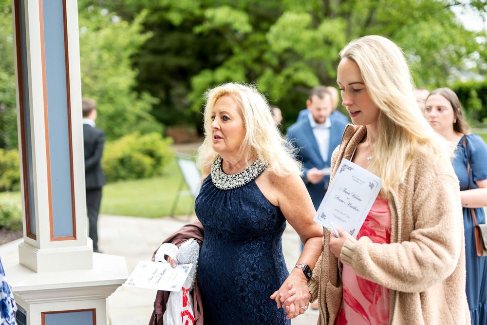Two elegantly dressed women holding wedding programs walk together at outdoor garden wedding venue.