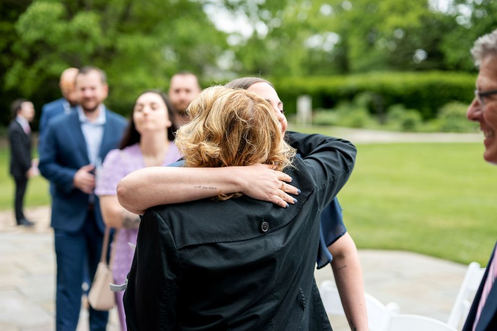 Two wedding guests share a warm embrace outdoors while other guests socialize in background.