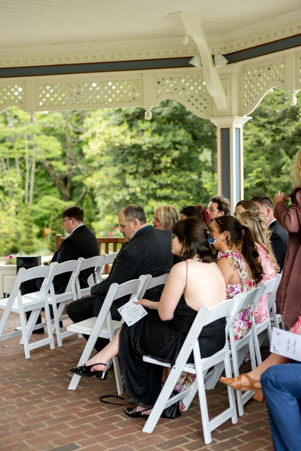 Wedding guests seated in white folding chairs under an elegant gazebo during an outdoor ceremony.