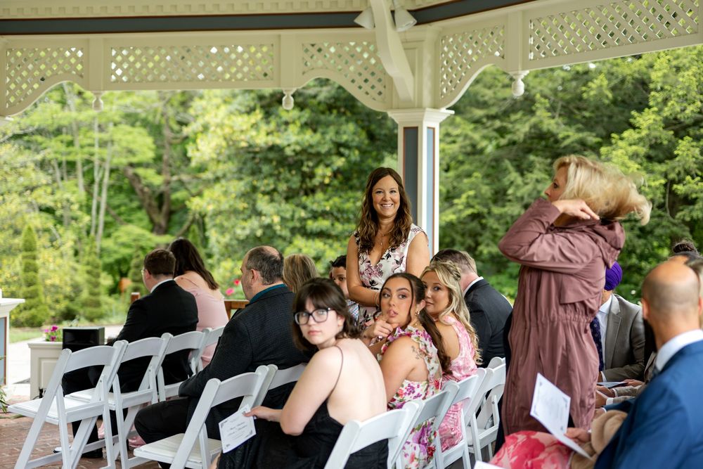 Wedding guests socialize under white gazebo at outdoor ceremony with lush green backdrop.