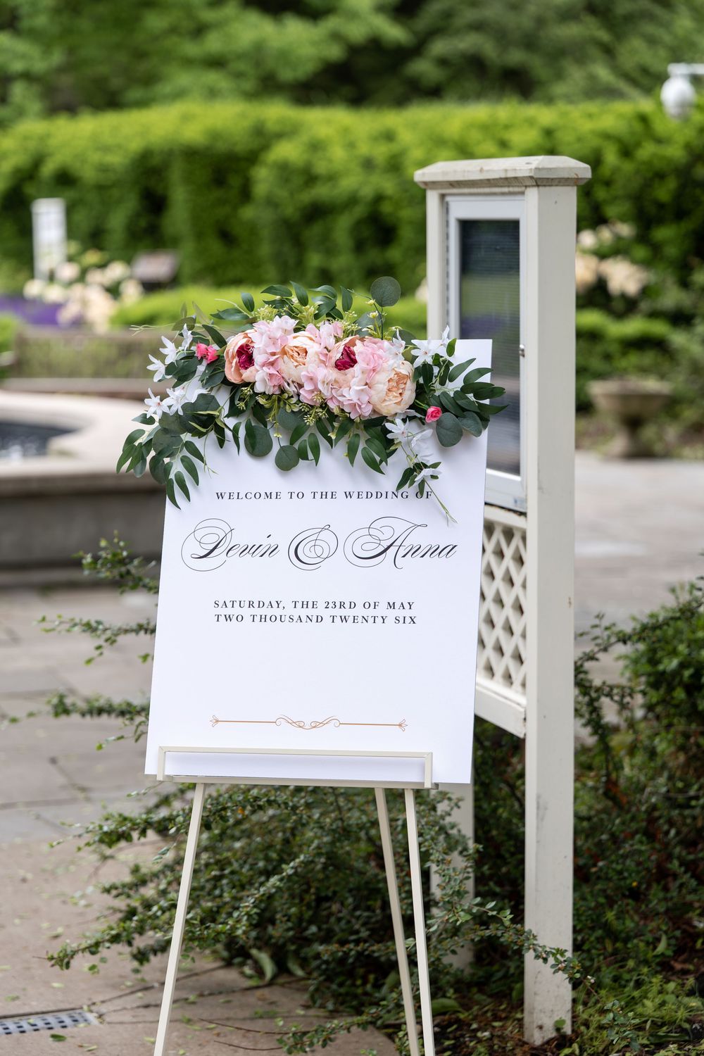 Elegant outdoor wedding welcome sign for Devin and Anna on Saturday May 23rd 2026 adorned with pink floral arrangement.