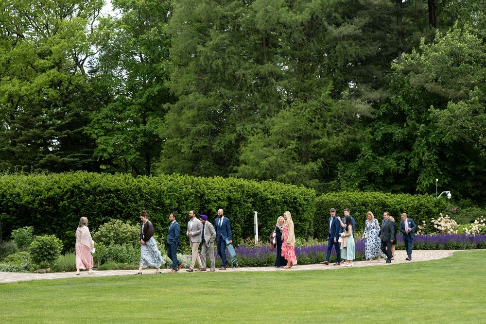 Wedding guests walk along a garden path lined with purple lavender and lush green hedges during an outdoor ceremony.