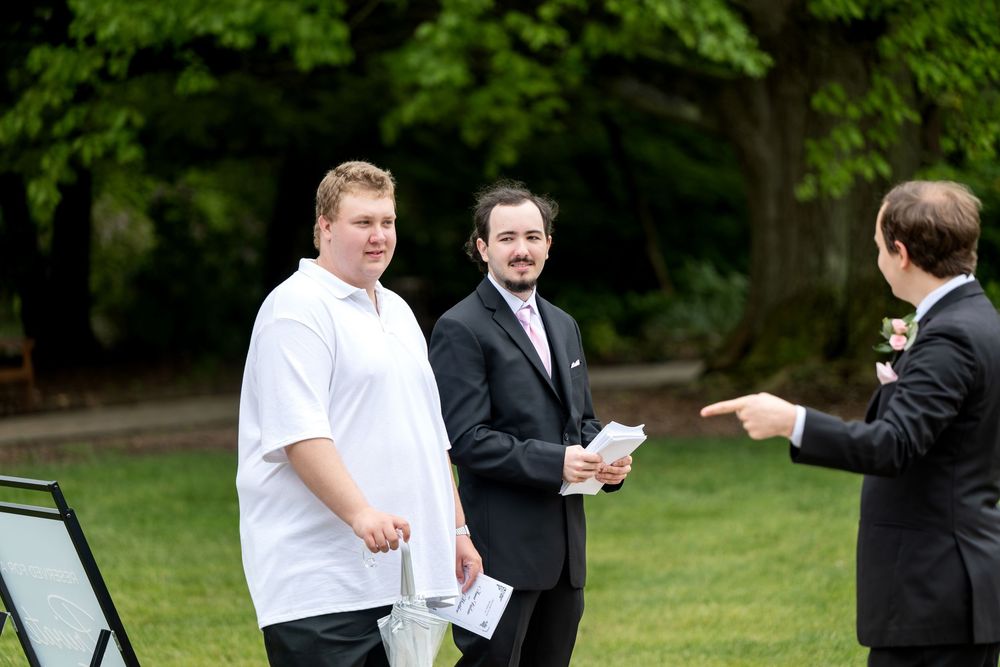 Three men in formal attire share a lighthearted moment outdoors at a wedding ceremony.