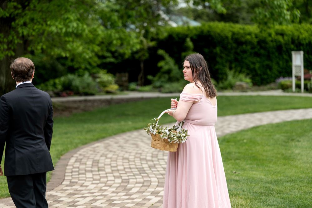 Bridesmaid in pink gown carries floral basket along brick garden path at outdoor wedding.