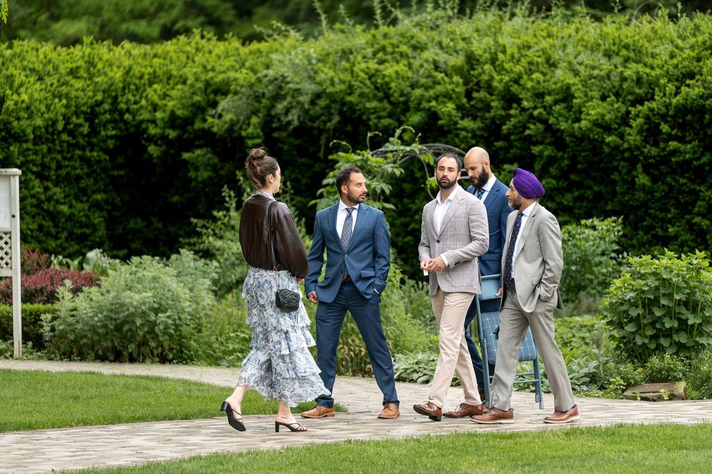 Wedding guests in formal attire walk along garden path surrounded by lush green hedges.