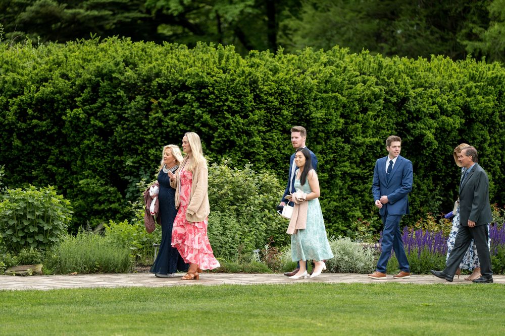 Wedding guests walking along garden path surrounded by lush green hedges and flowers.