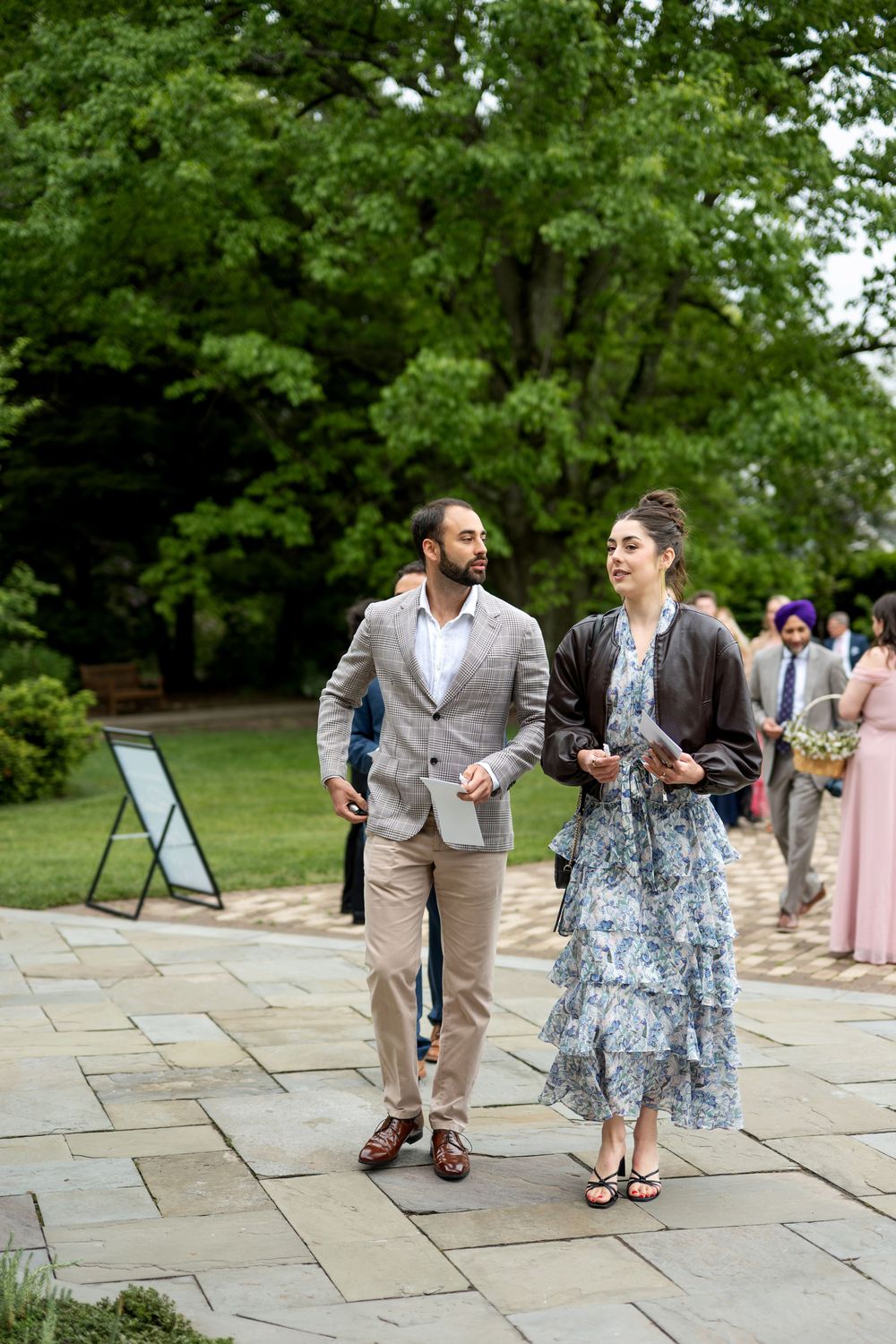 Wedding guests in formal attire walk along stone pathway surrounded by lush green gardens.
