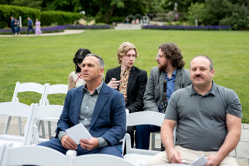 Wedding ceremony guests seated on white chairs outdoors on lush green lawn holding programs.