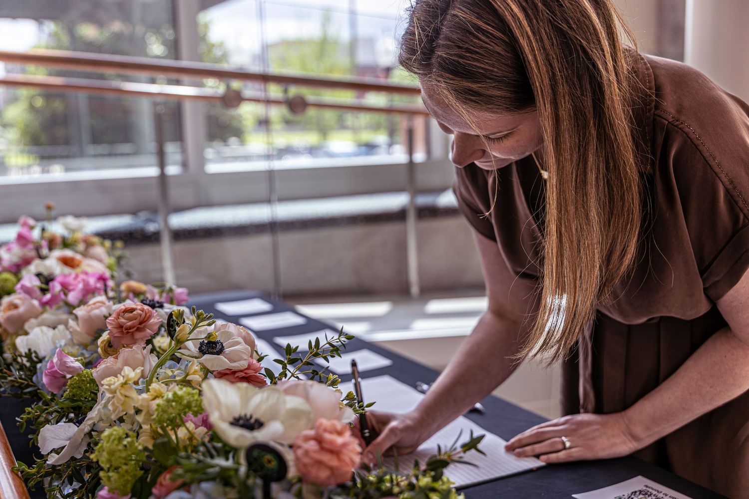 Woman in brown dress signing documents beside elegant floral arrangements of pink and white flowers at an event.