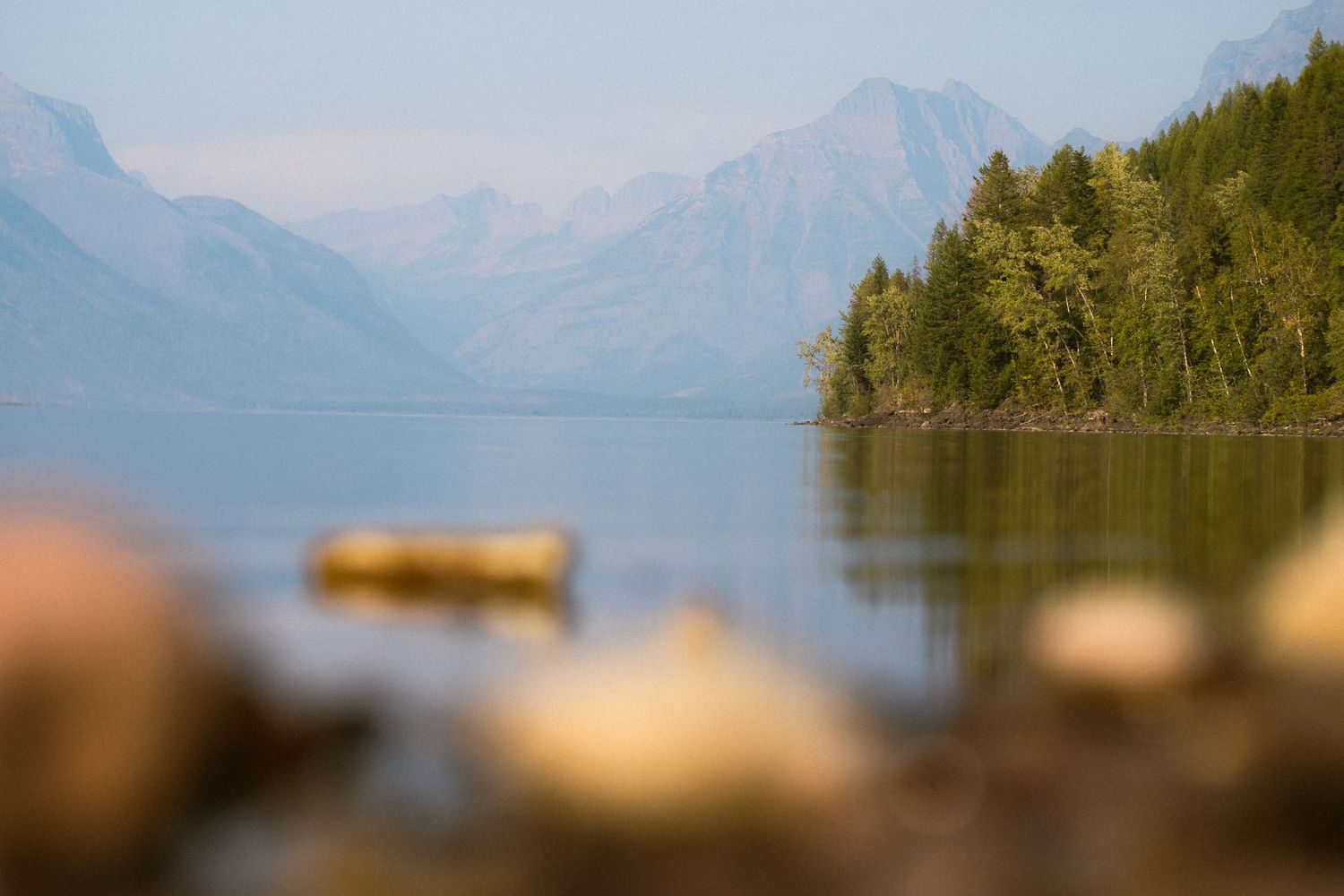Serene McDonald lake in Glacier with misty peaks and forested shoreline reflected in calm blue water.