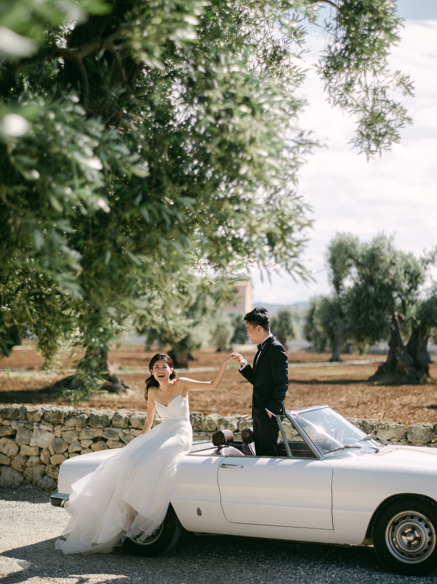 Bride and groom posing with a white vintage convertible car among olive trees at an outdoor wedding venue.